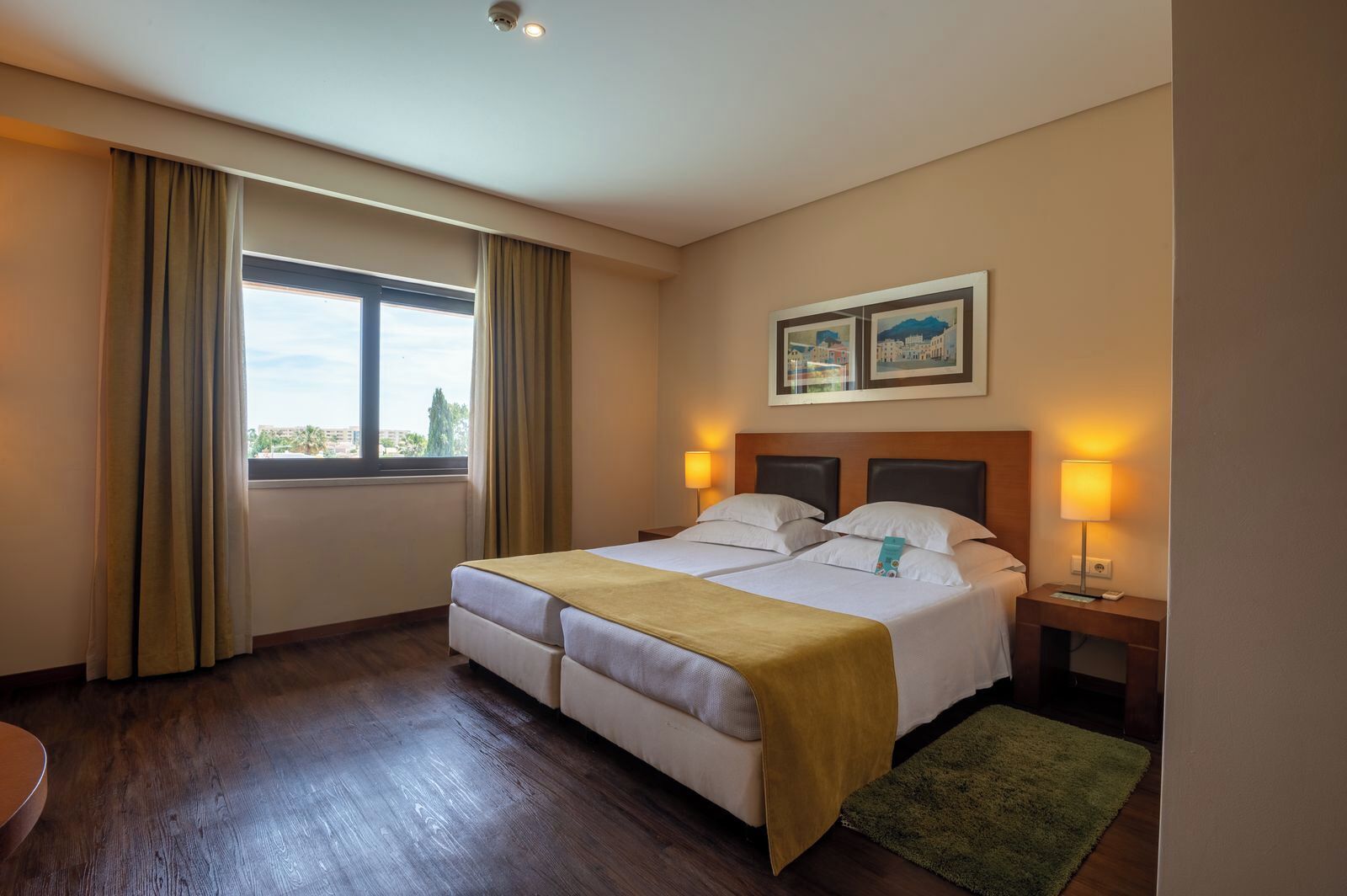 Habitación de hotel con una cama doble, dos lámparas de noche, una ventana con cortinas, un cuadro enmarcado en la pared y un escritorio pequeño con una lámpara.