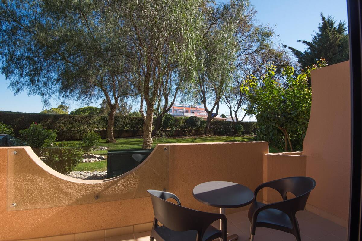 A balcony with two chairs and a table, overlooking a landscaped garden with trees and a building in the background.
