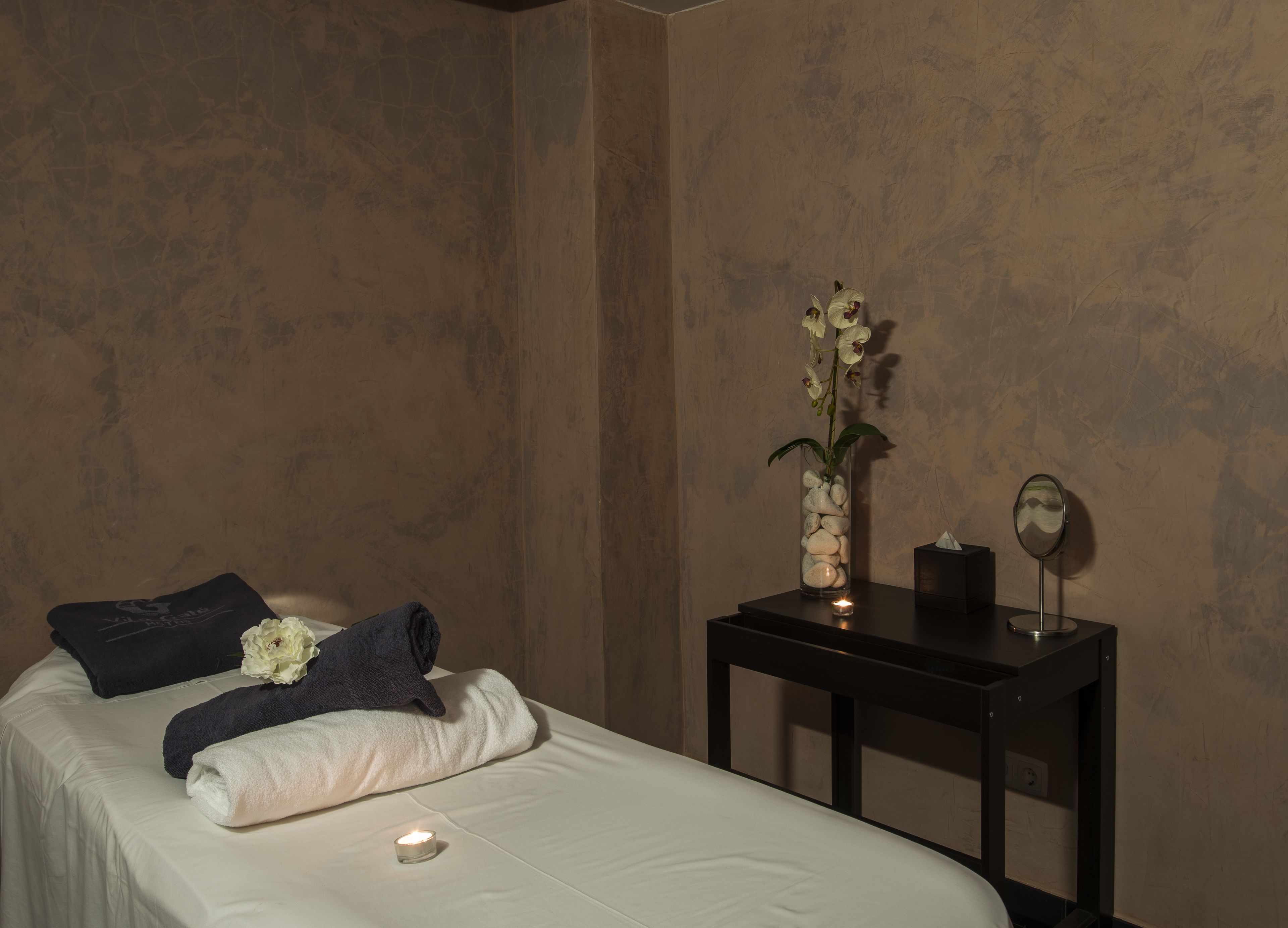 Una cama bien hecha con toallas, un florero con flores, una vela y un espejo en una mesita de noche en una habitación con paredes texturizadas.