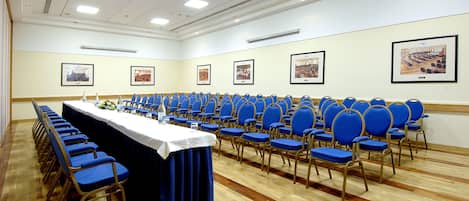 Un salón de conferencias con filas de sillas azules, una larga mesa con mantel blanco y cuadros en las paredes.