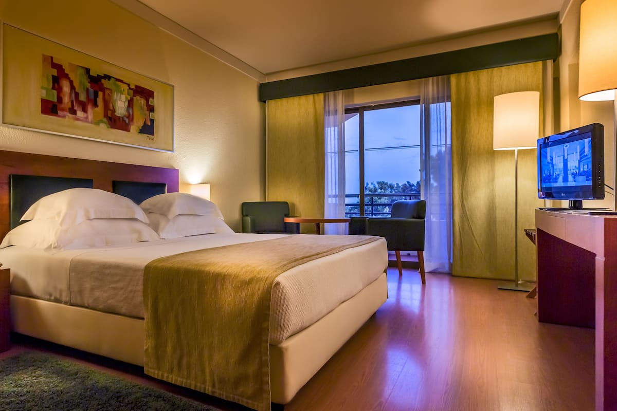 A hotel room with a bed, bedside tables, a desk with a chair, a TV, and a large window with curtains.