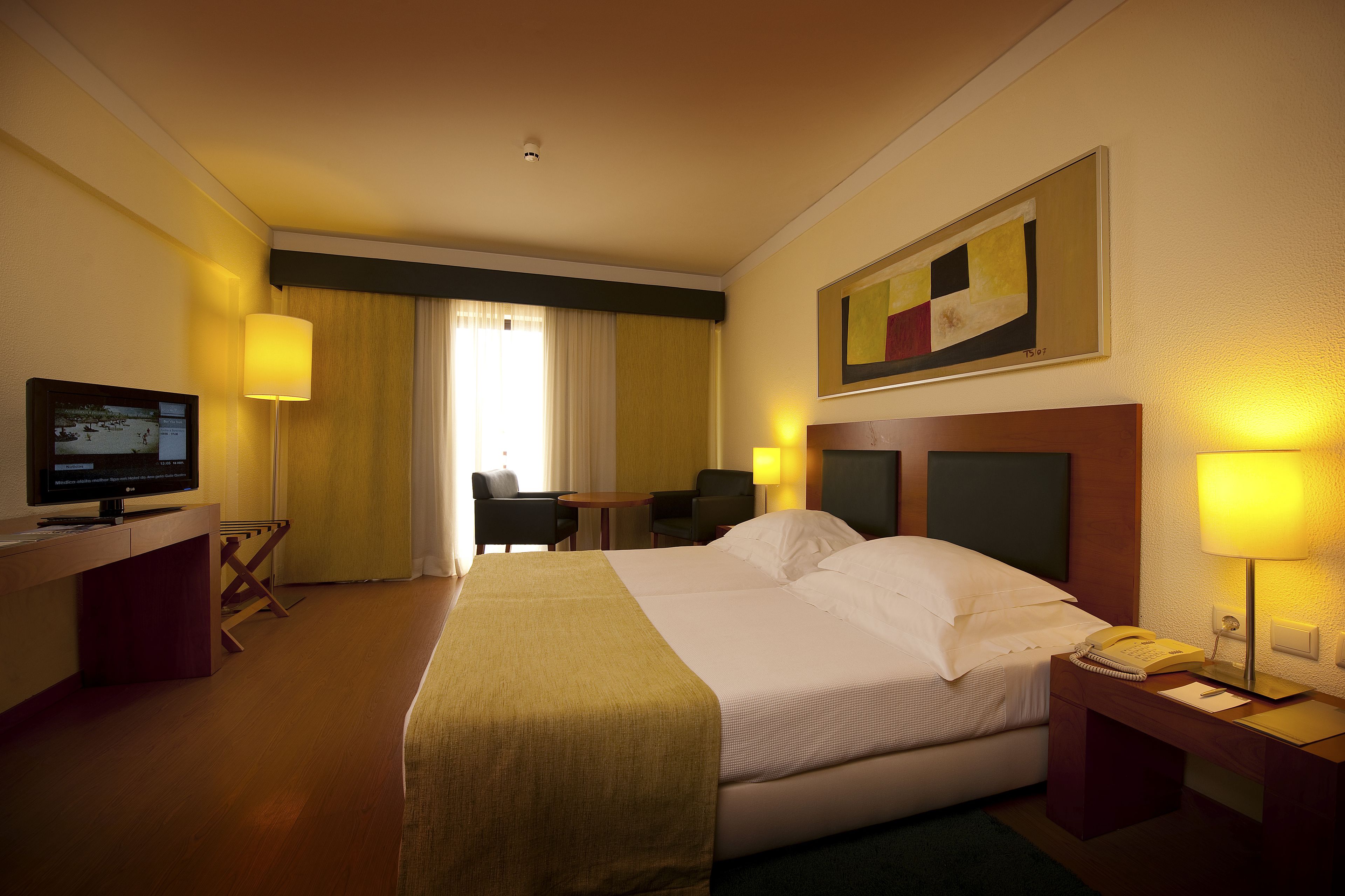 A hotel room with a bed, bedside table, lamp, desk, chair, TV, and a painting on the wall.