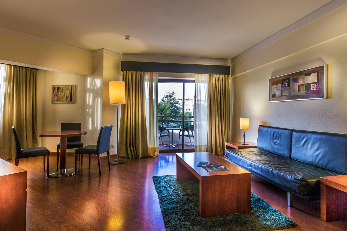 Een hotelkamer met een houten bureau, een bank, een eettafel met stoelen en uitzicht op het buitenleven door een groot raam.