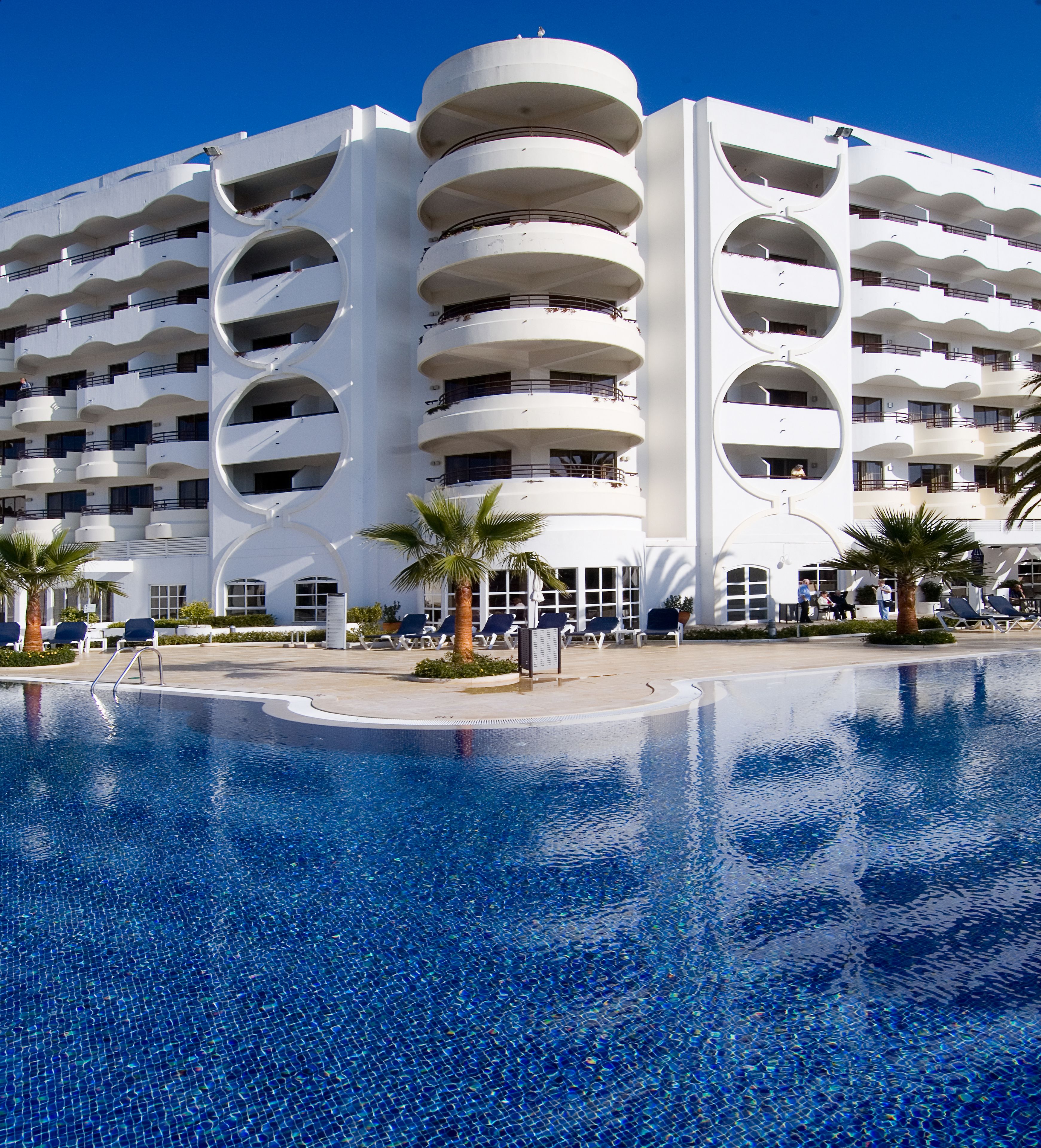 Ein modernes Hotel mit großem Pool, mehreren Balkonen und Palmen.
