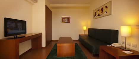 A hotel room with a wooden desk, a television, a sofa, a green rug, and two bedside lamps.