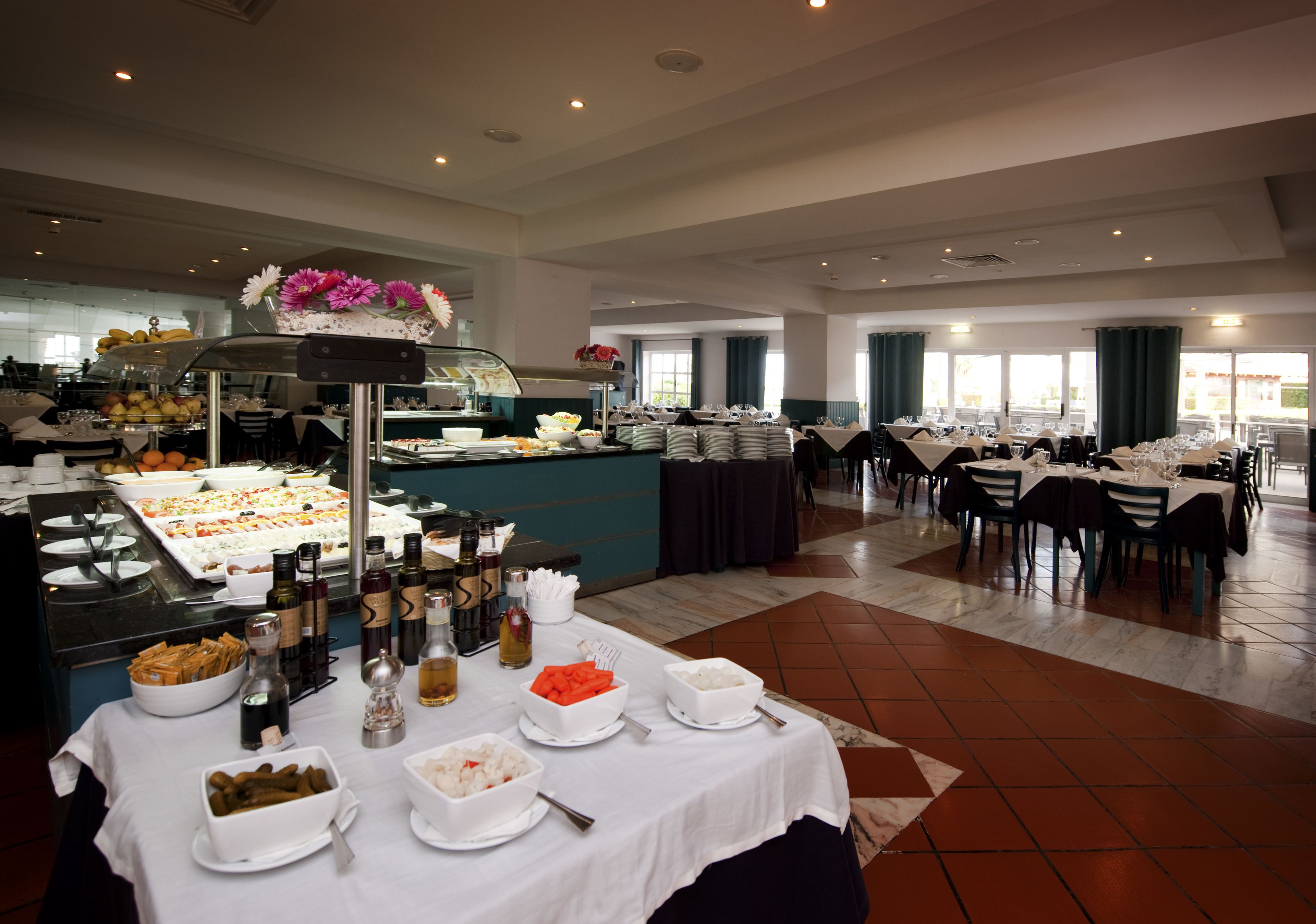 Un buffet proposant une variété de plats et de boissons, des tables dressées pour le repas et une grande fenêtre laissant entrer la lumière naturelle.