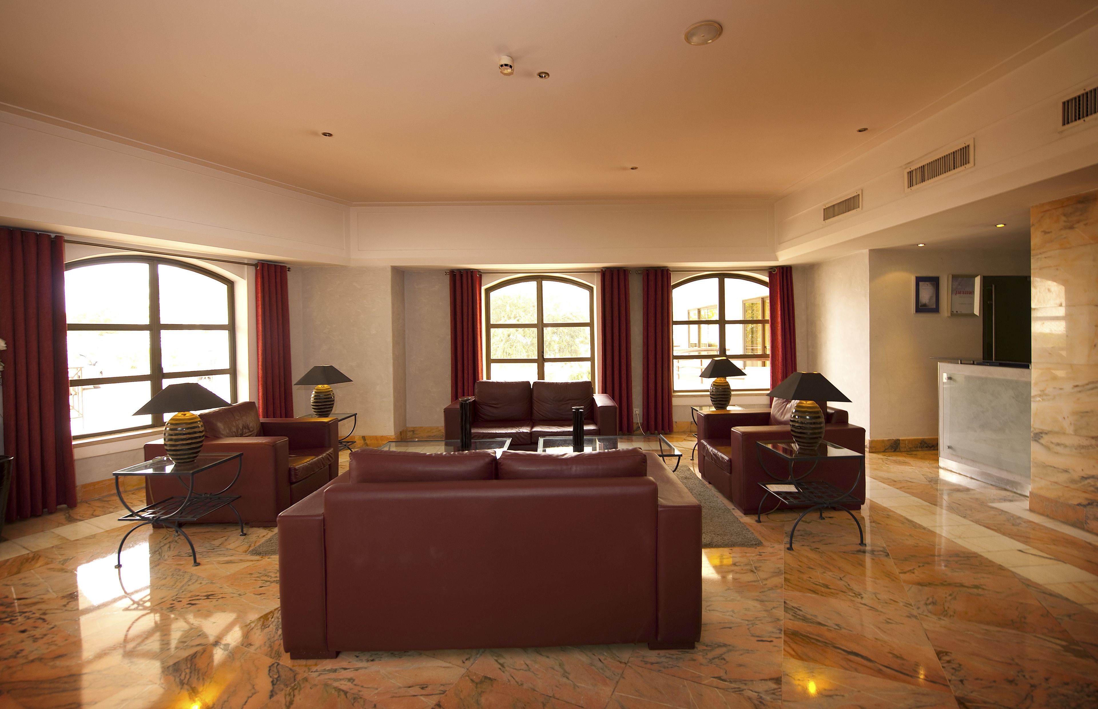 Uma sala de estar ampla com um sofá marrom grande, duas poltronas e uma mesa de centro. Há janelas grandes com cortinas vermelhas, uma lareira e um piso de mármore.