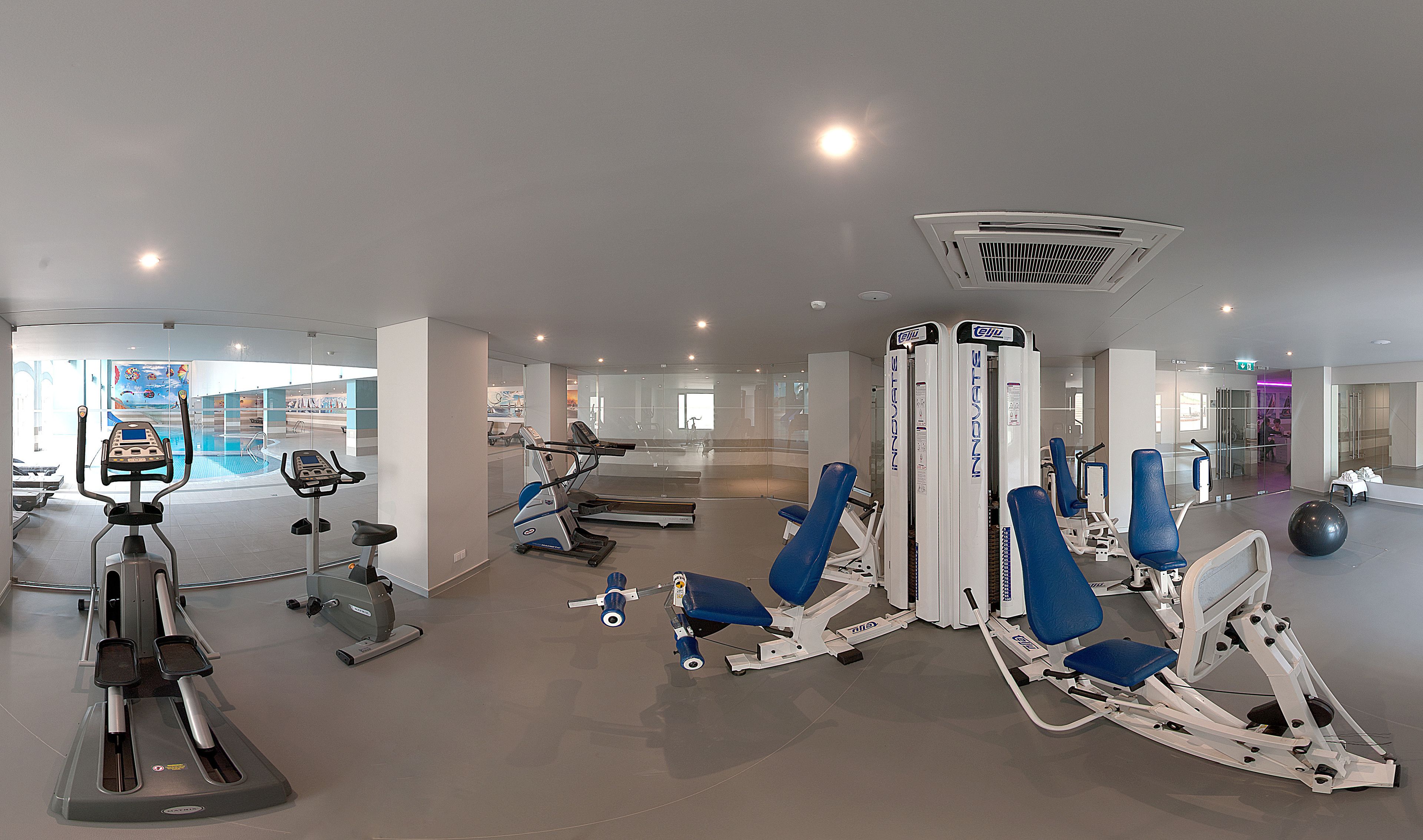 A modern gym with various exercise equipment, a mirrored wall, and a pool area visible in the background.