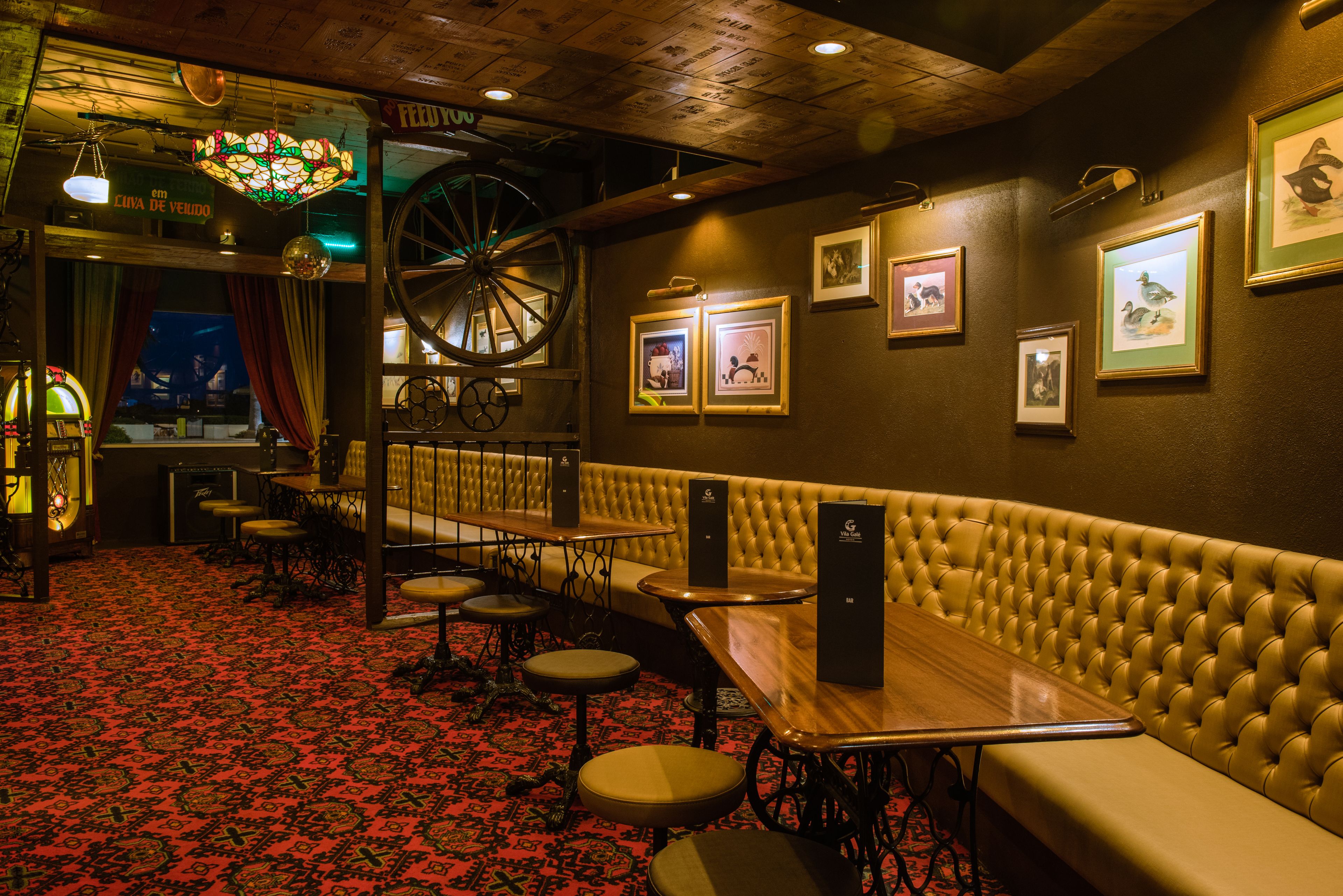A cozy interior with a red patterned carpet, a wooden bar area, and a seating section with a tufted bench and tables.