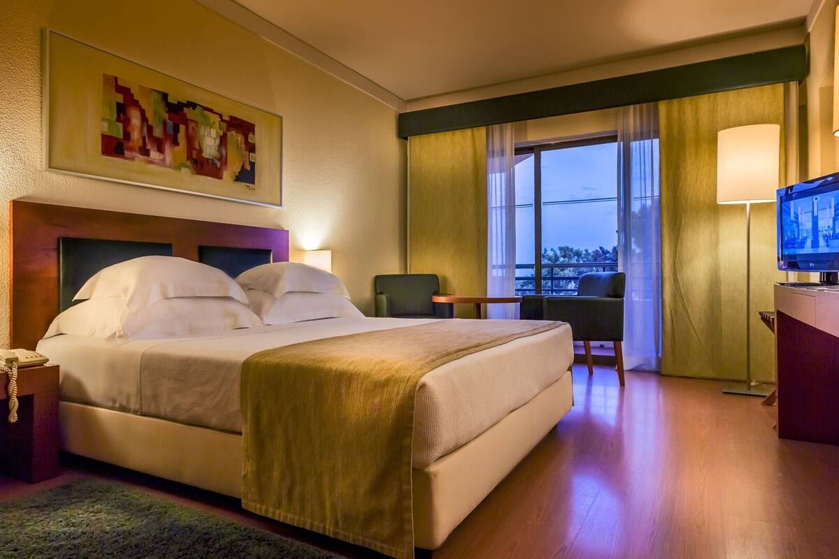A hotel room with a bed, bedside tables, a desk, a chair, a TV, and a large window.