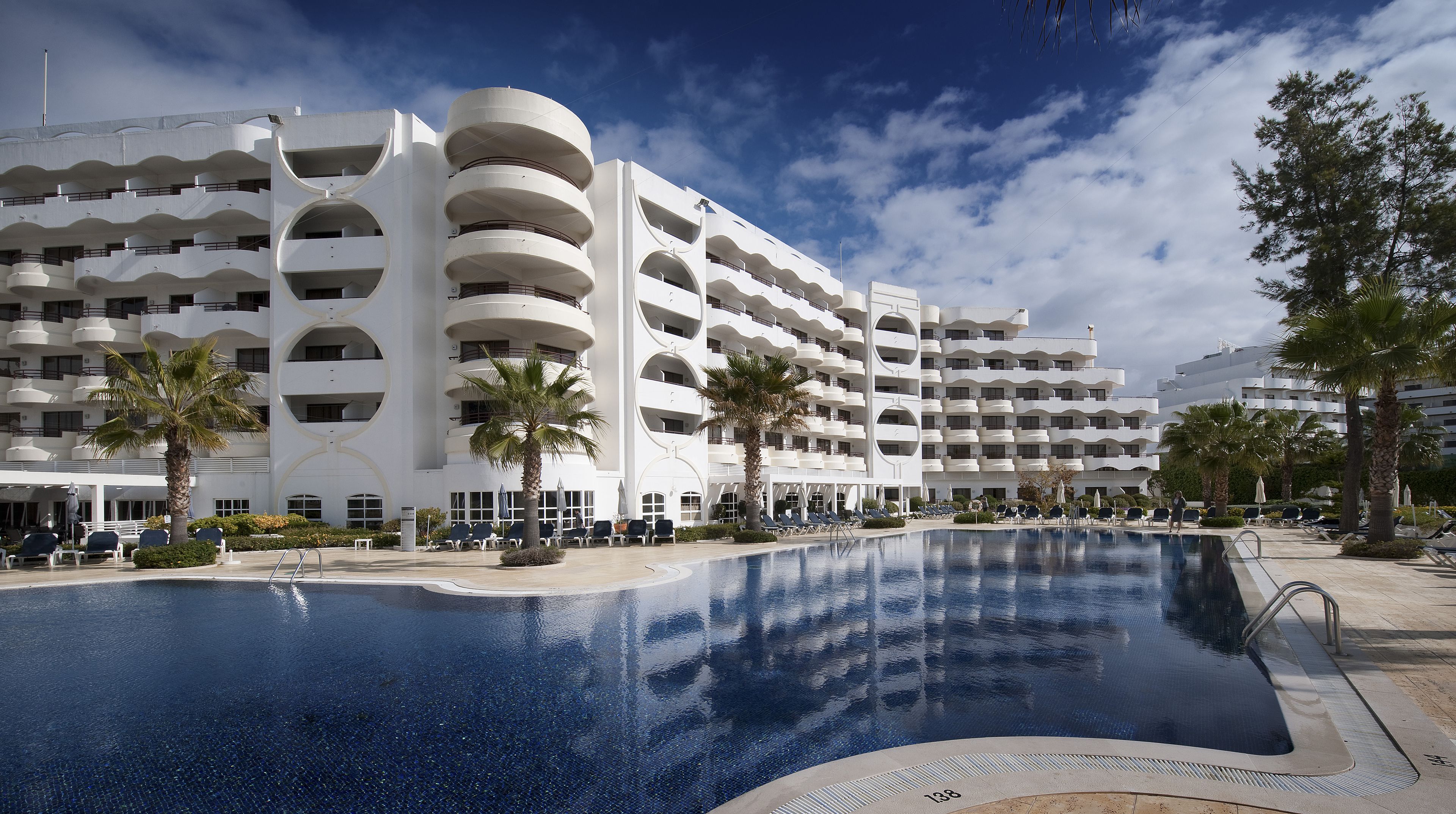 Un hôtel moderne doté d’une grande piscine, de nombreux balcons et de palmiers.