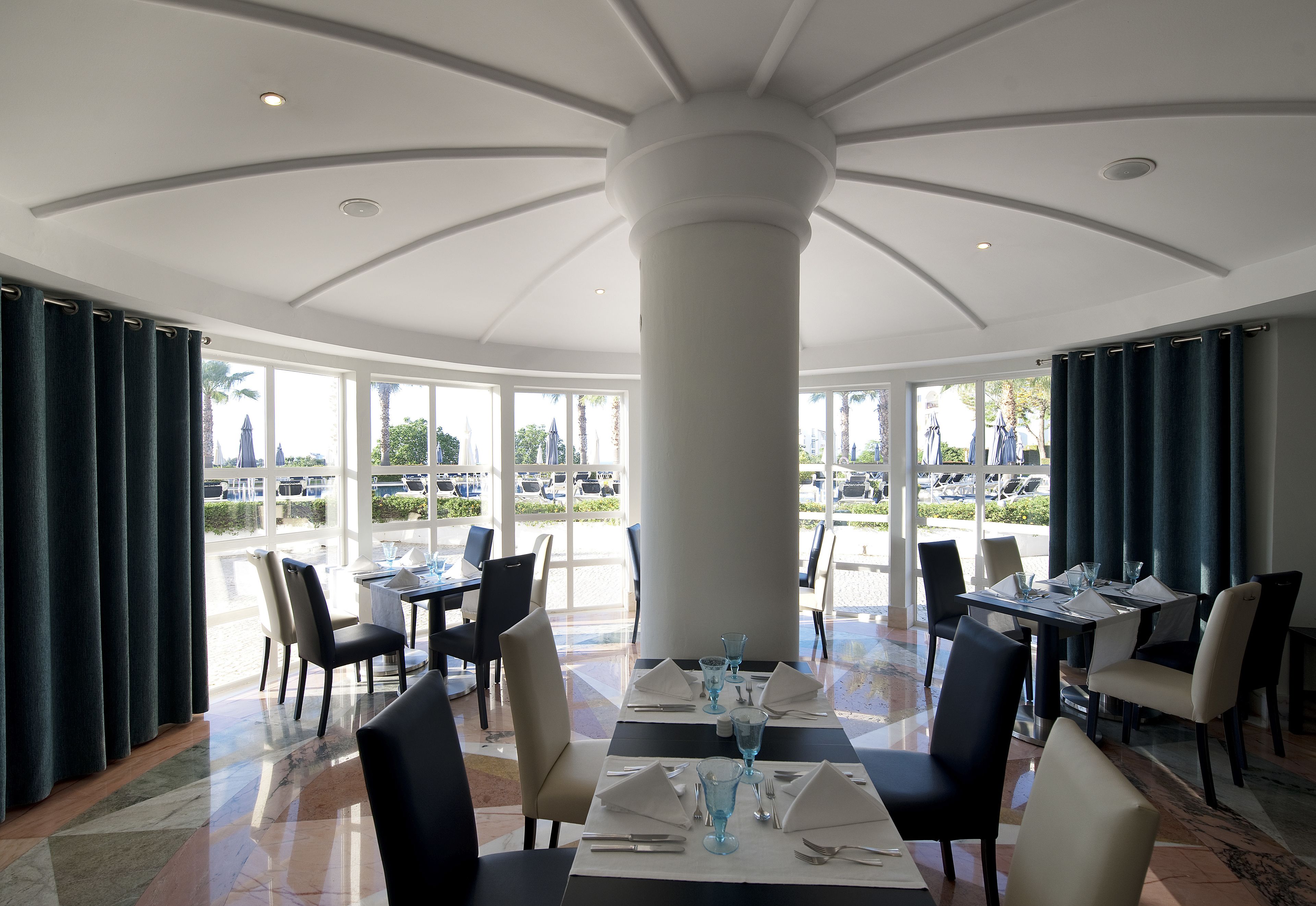 A dining area with tables set for four, large windows, and a central pillar.