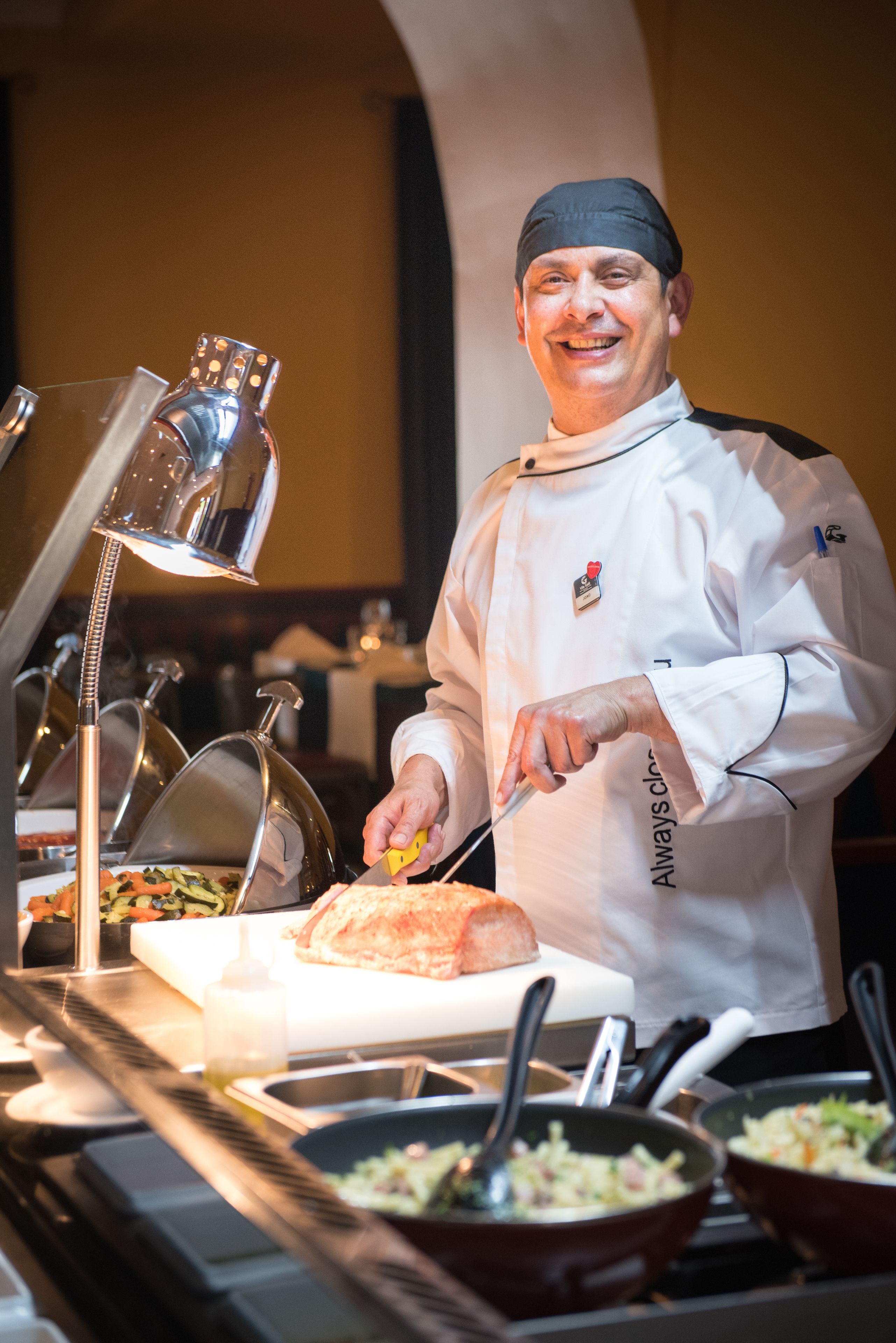 Un chef con uniforme blanco y gorro negro preparando comida en una estación de buffet con diversos platos y utensilios de cocina.