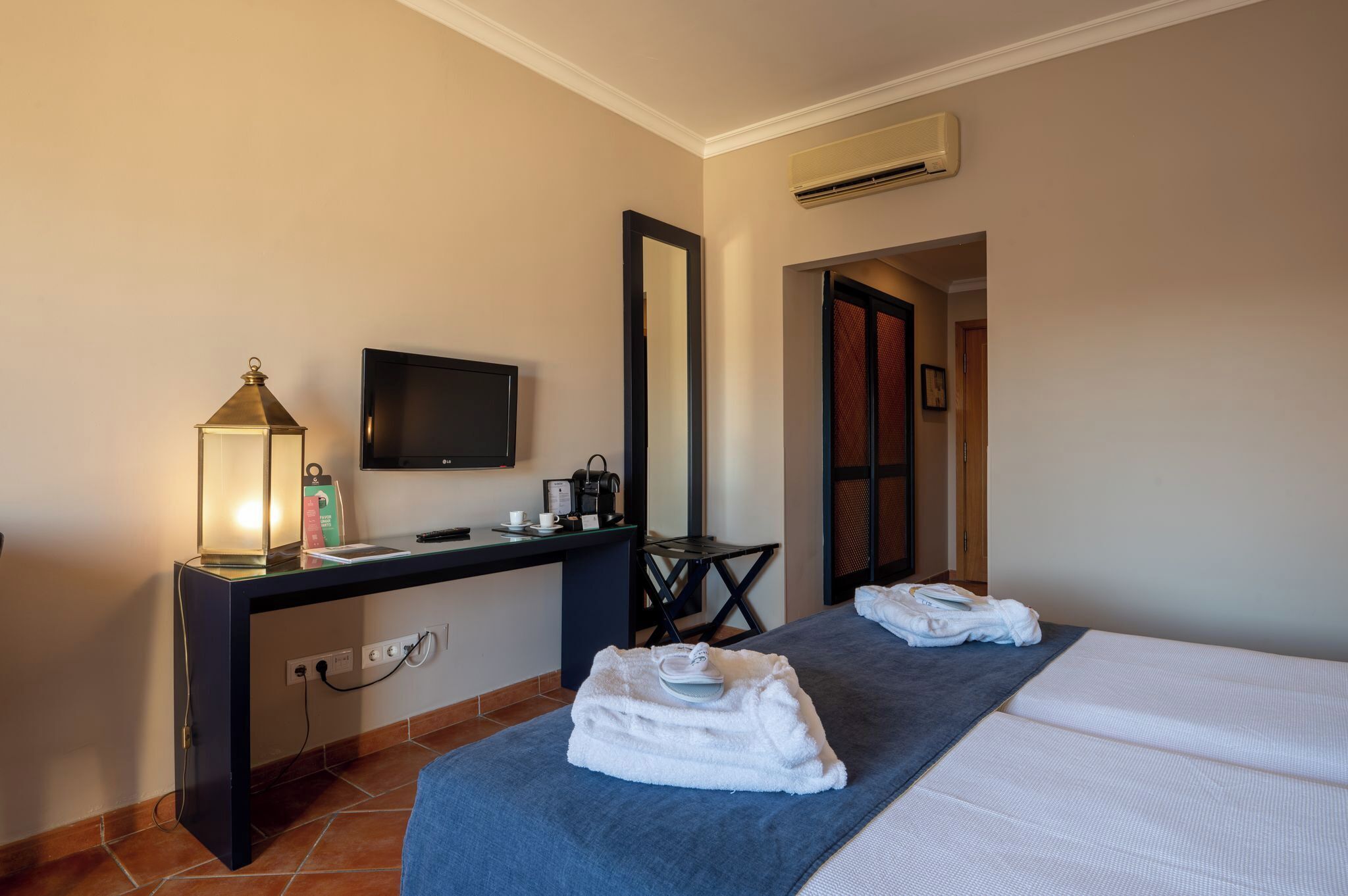 A hotel room with a bed, a desk with a TV, a lamp, and a mirror.