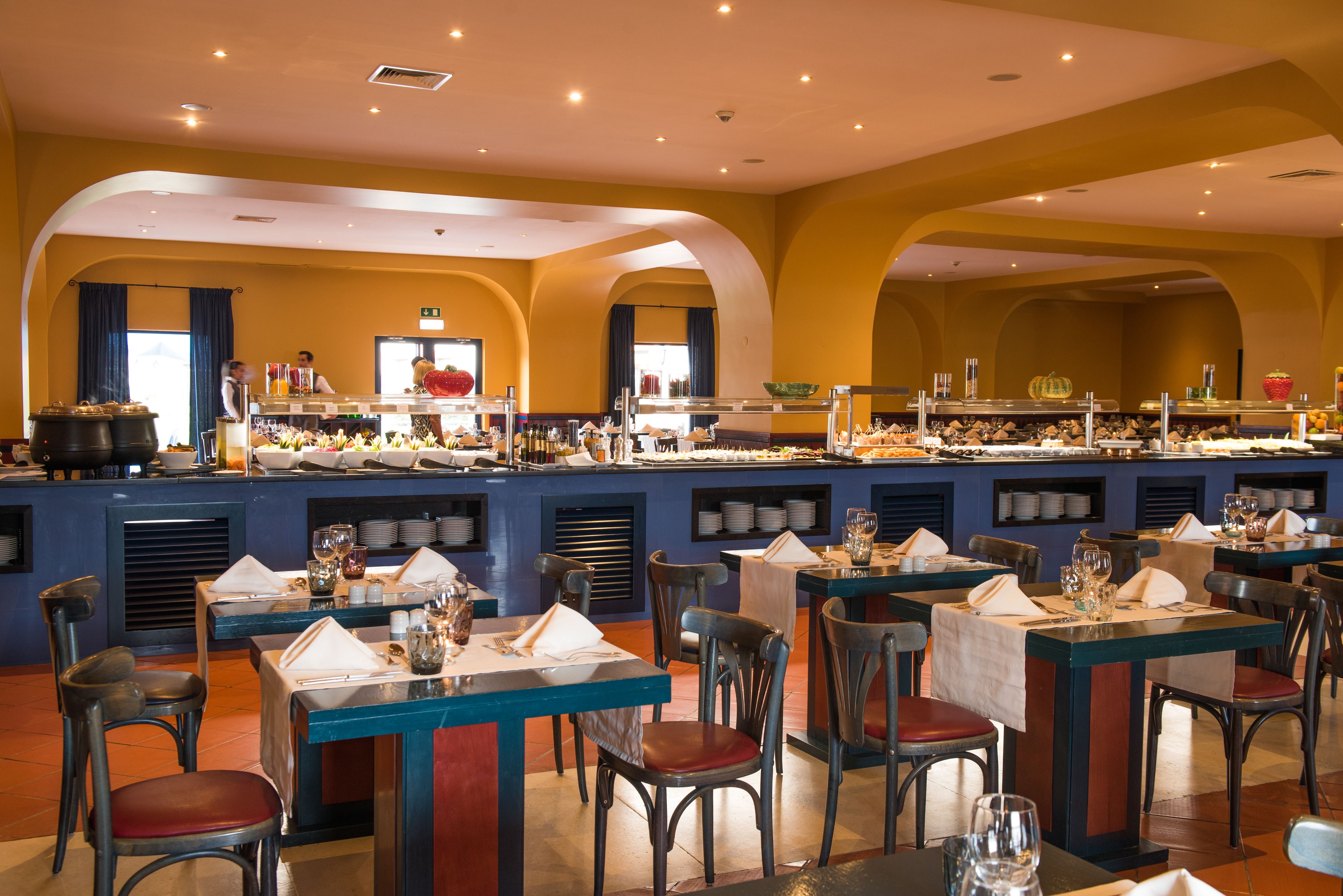 Ein Restaurant im Buffetstil mit mehreren Tischen und Stühlen, einer zentralen Servicestation und einer Farbgestaltung in Gelb und Blau.