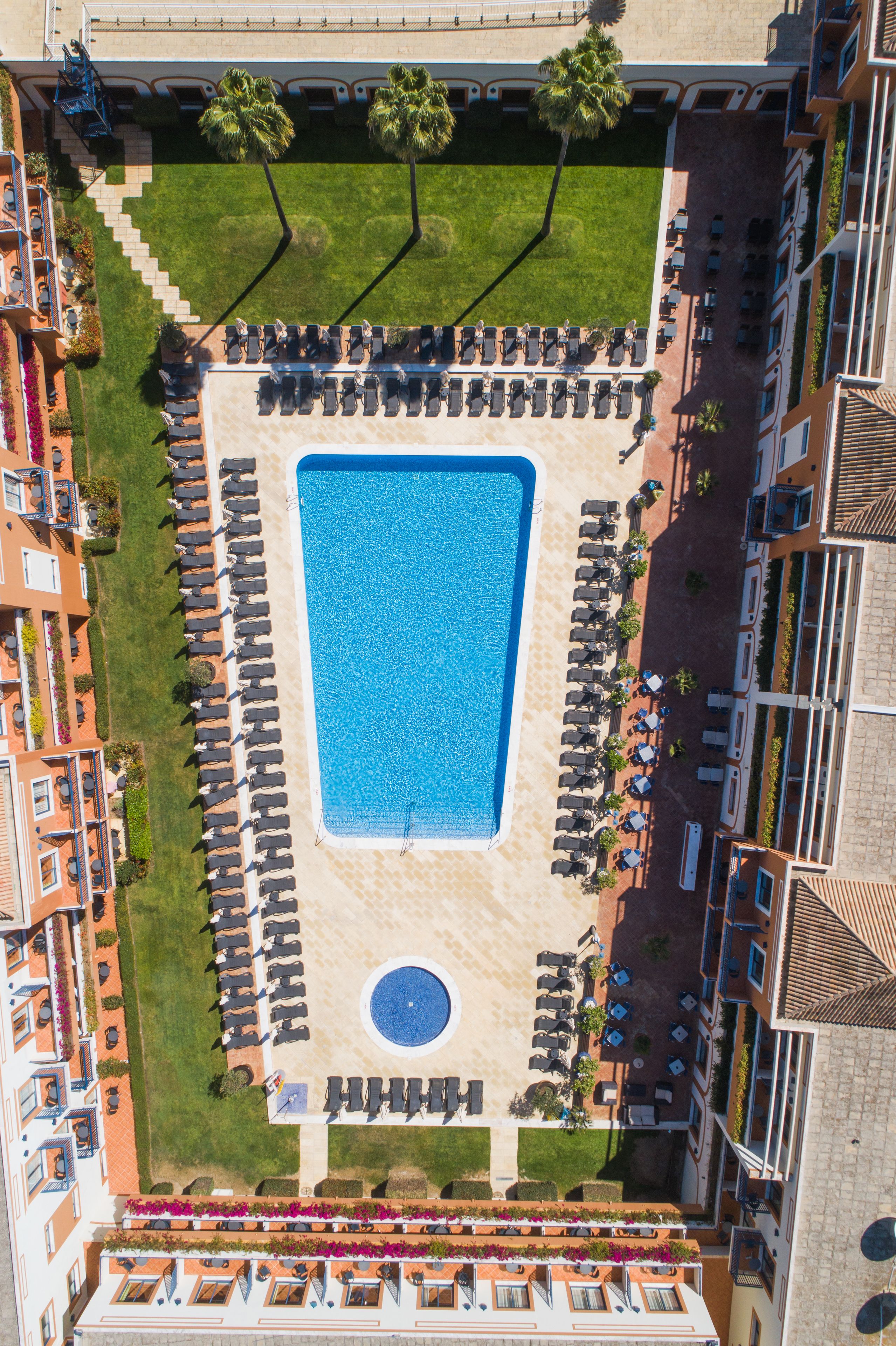 Vista aérea de un hotel con piscina central, sillones de descanso y jardín ajardinado.