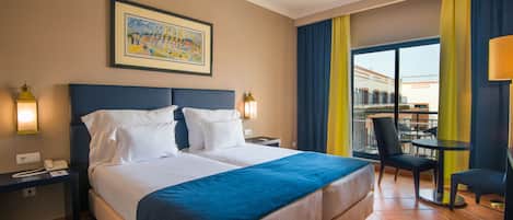 A hotel room with a large bed, a desk with a lamp, a chair, and a balcony with a view of buildings.