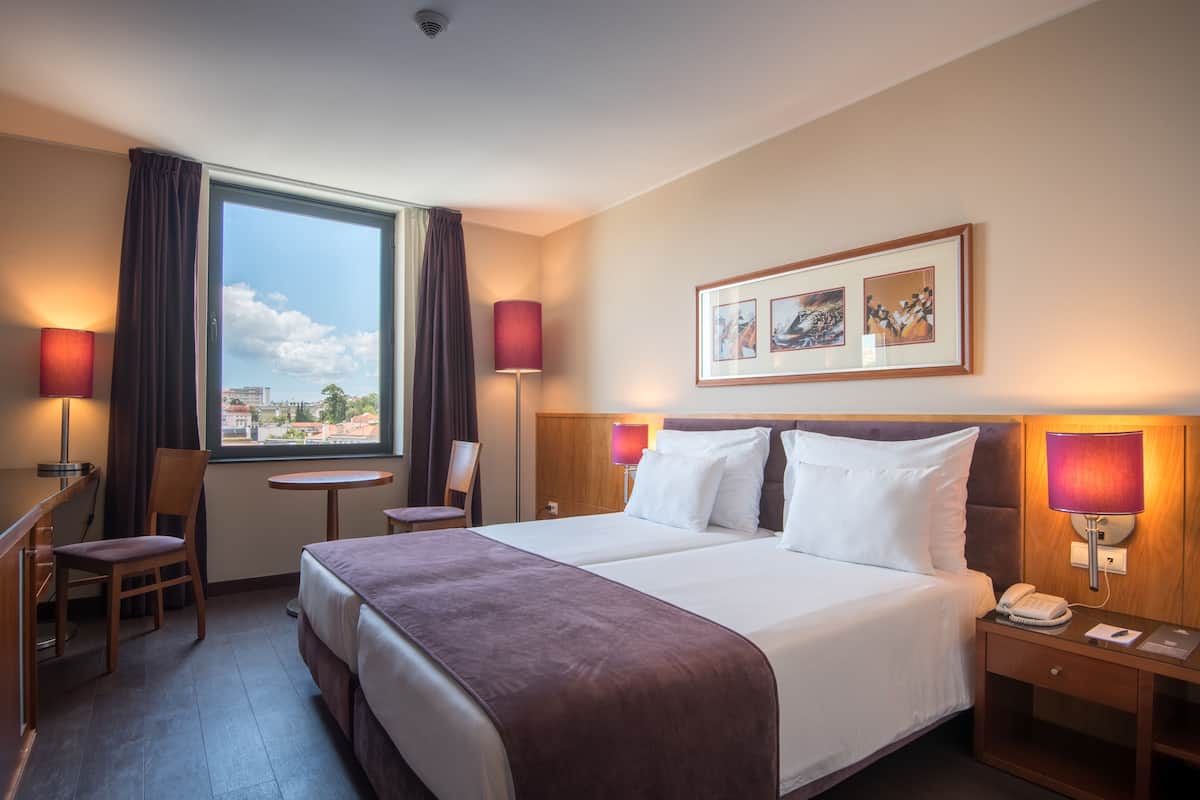 A hotel room with a large bed, two bedside lamps, a desk with a chair, a small table, and a window with curtains.