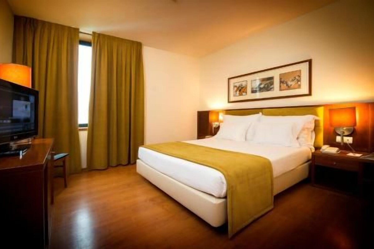 A hotel room with a bed, a television, a desk, and a window with curtains.