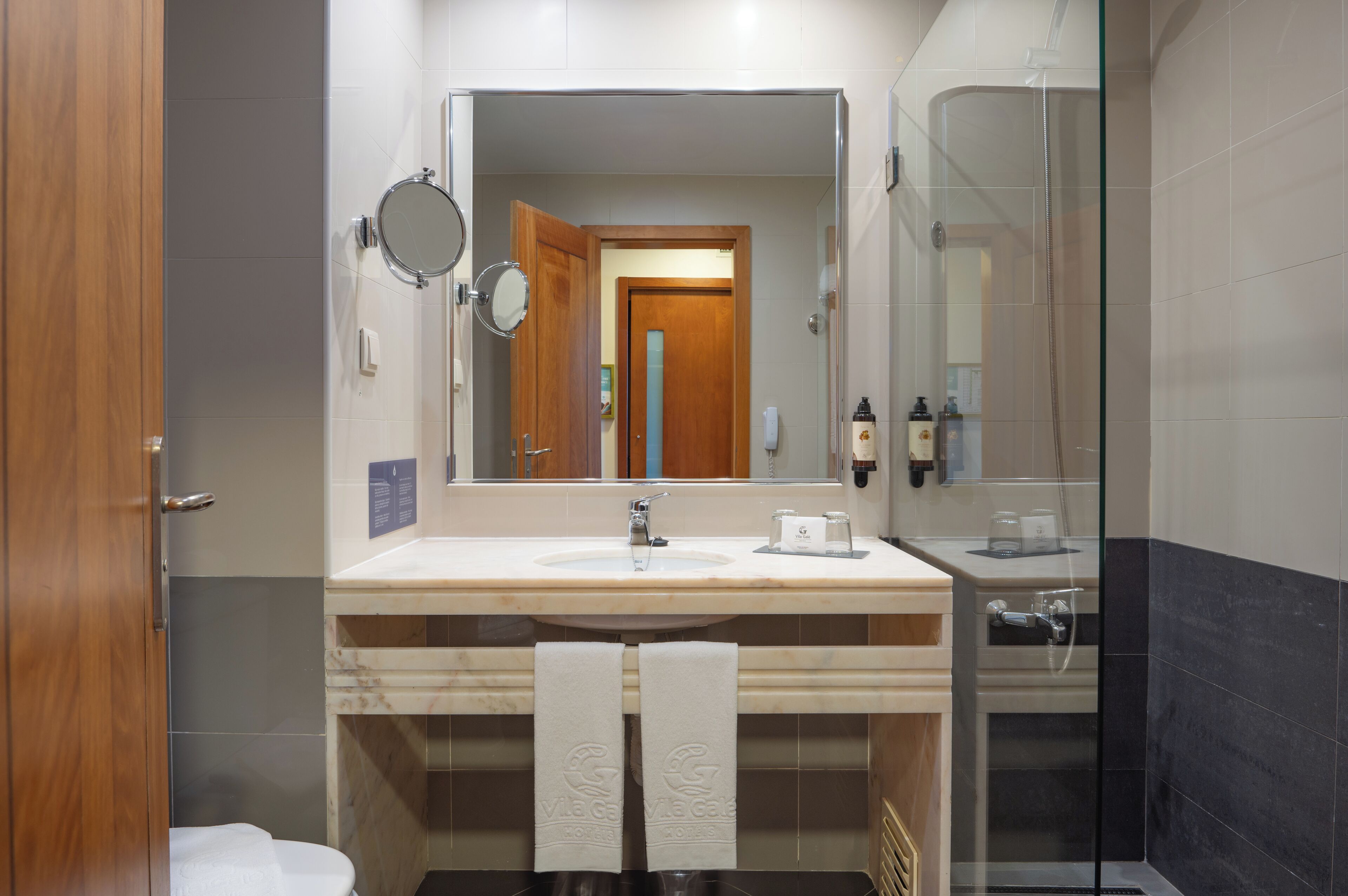 A bathroom with a large mirror, a sink, a glass shower enclosure, and two towels hanging on the wall.
