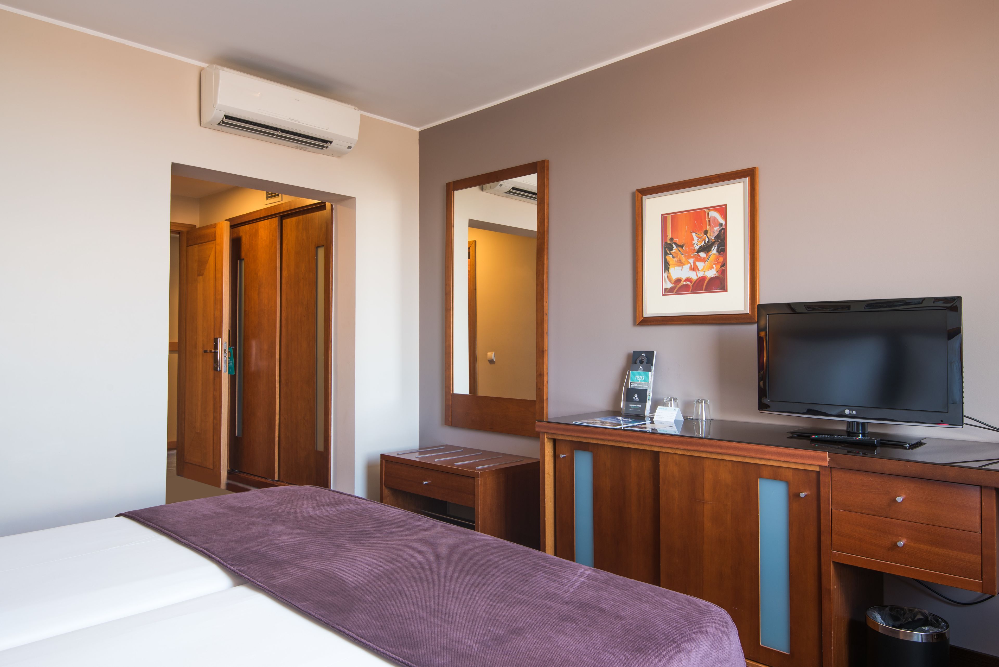 A hotel room with a bed, a desk, a television, a mirror, and an air conditioning unit.