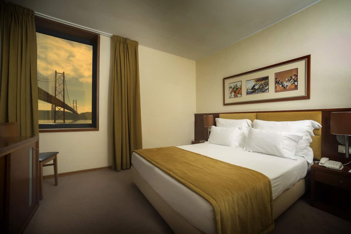 A hotel room with a large bed, a desk, a chair, a window with a view of a bridge, and framed artwork on the wall.