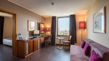 Ein Hotelzimmer mit Bett, Schreibtisch mit Computer, einem kleinen Tisch mit Stühlen, einer Couch und einem großen Fenster mit Vorhängen.