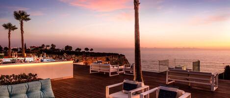 Um deck de madeira com móveis de exterior brancos, área de bar e vista para o oceano ao pôr do sol.