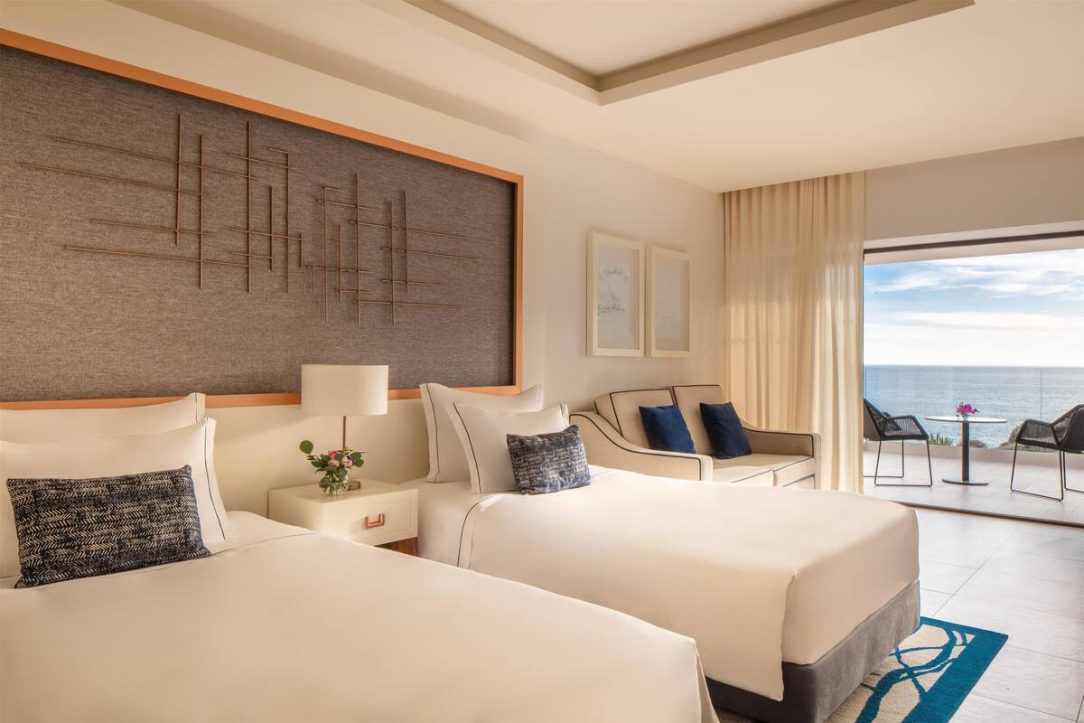A hotel room with two beds, a large window with a view of the sea, a balcony with chairs, and a wall with abstract art.
