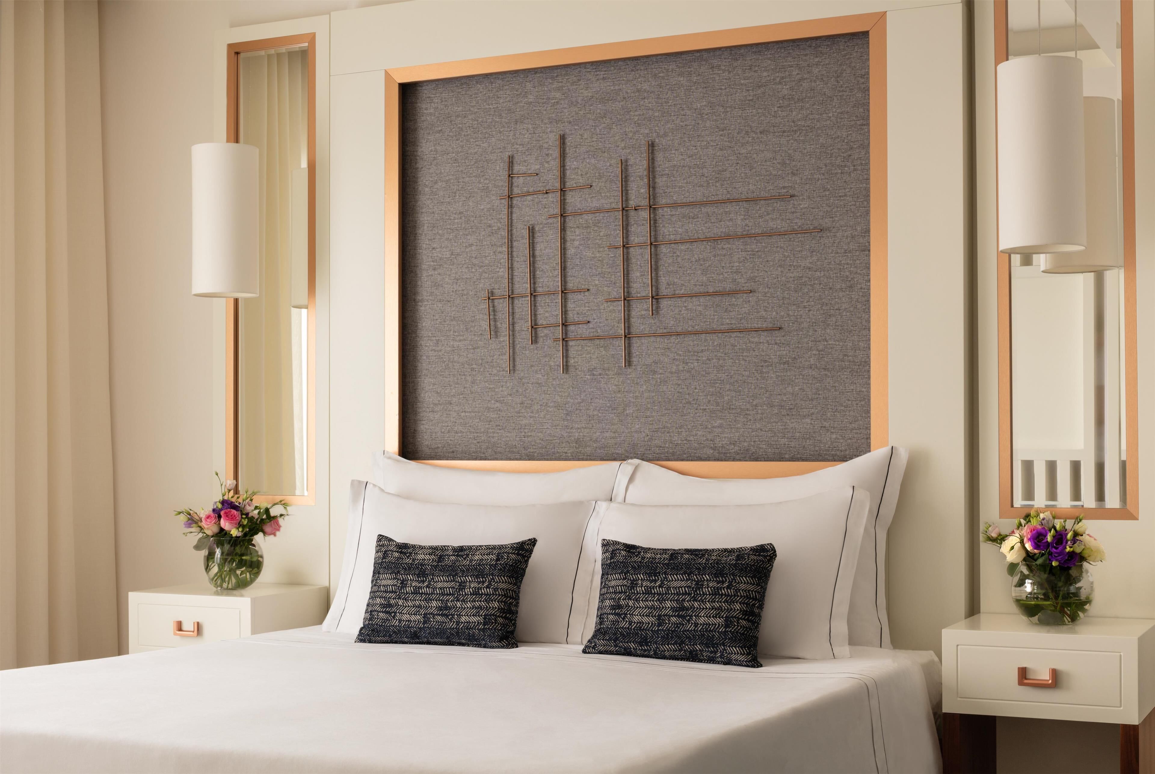 A modern hotel room with a large bed, two bedside tables with vases of flowers, a large artwork on the wall, and a large mirror.