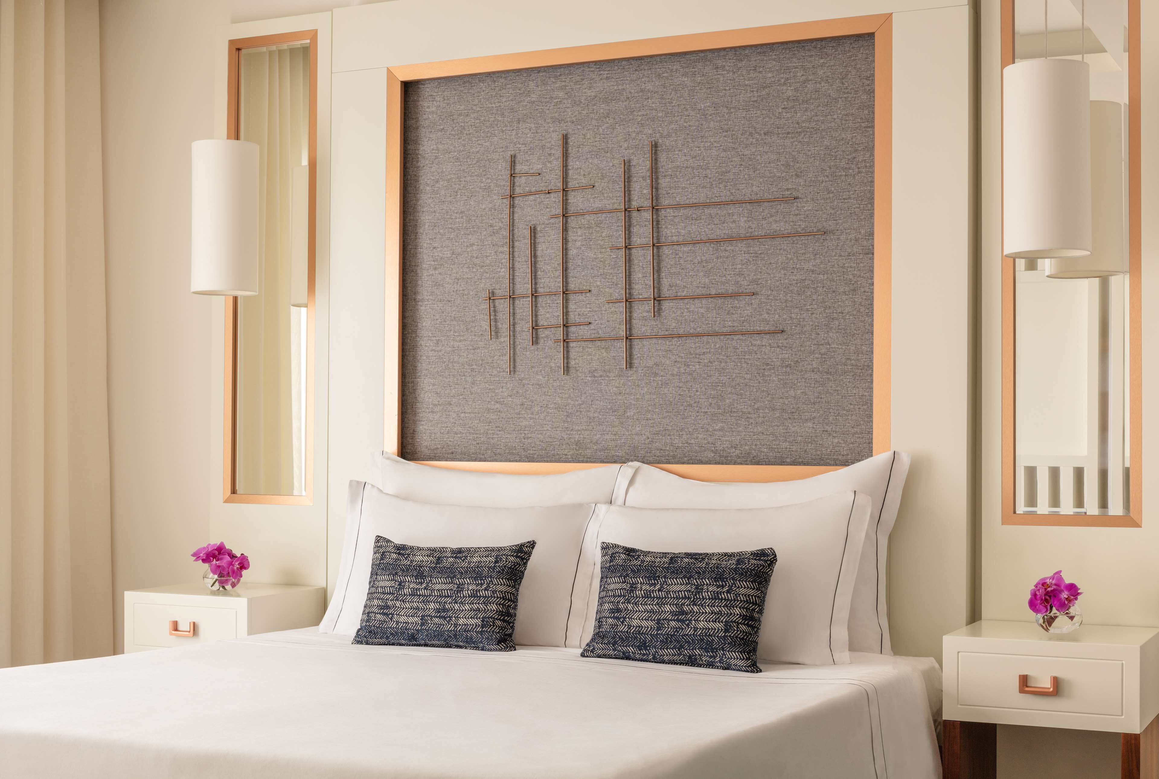 A modern hotel room with a large bed, two decorative pillows, a wall-mounted artwork, and a bedside table with a vase of flowers.