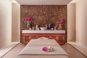 A spa room with a massage table, a wooden cabinet with decorative items, and a marble wall.