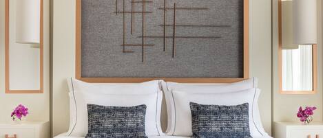 A hotel room with a large bed, two decorative pillows, a grey textured wall art, and a wooden headboard.
