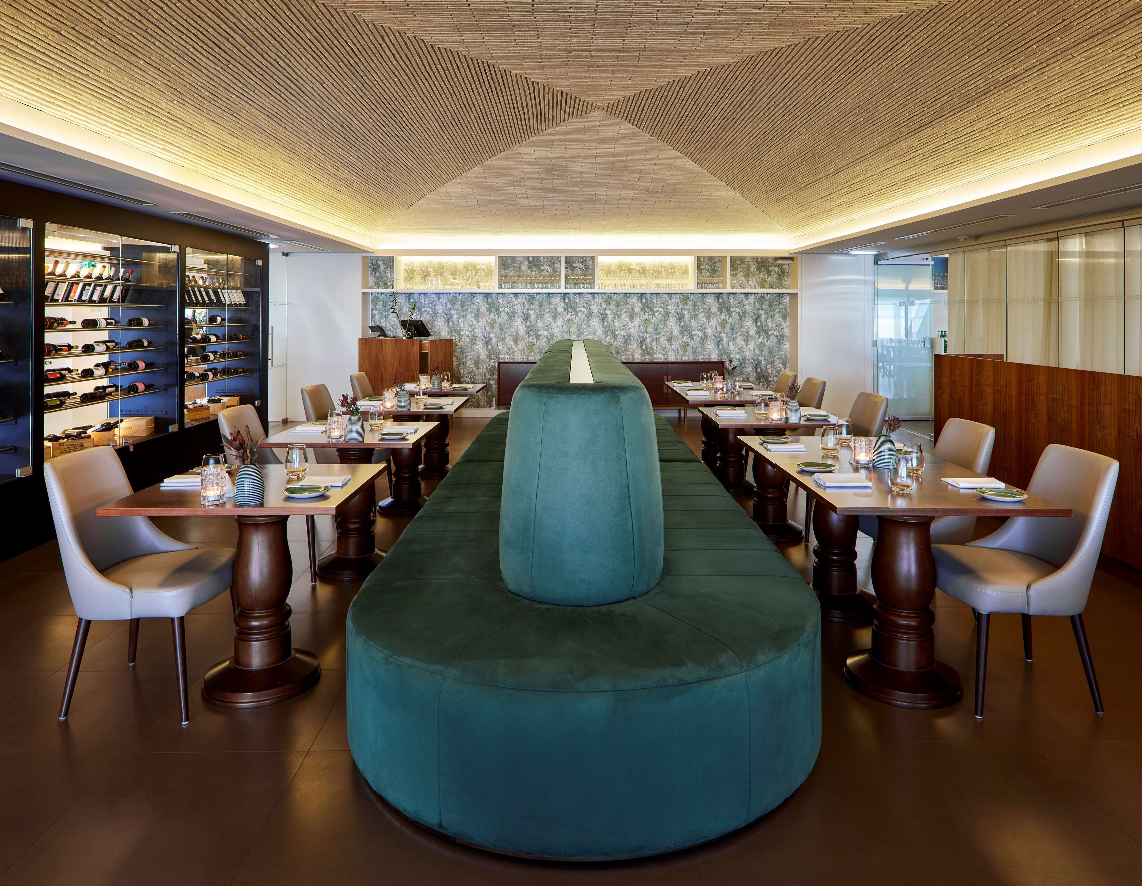 A restaurant with a long table, a green cushioned bench, and wine racks on the left.