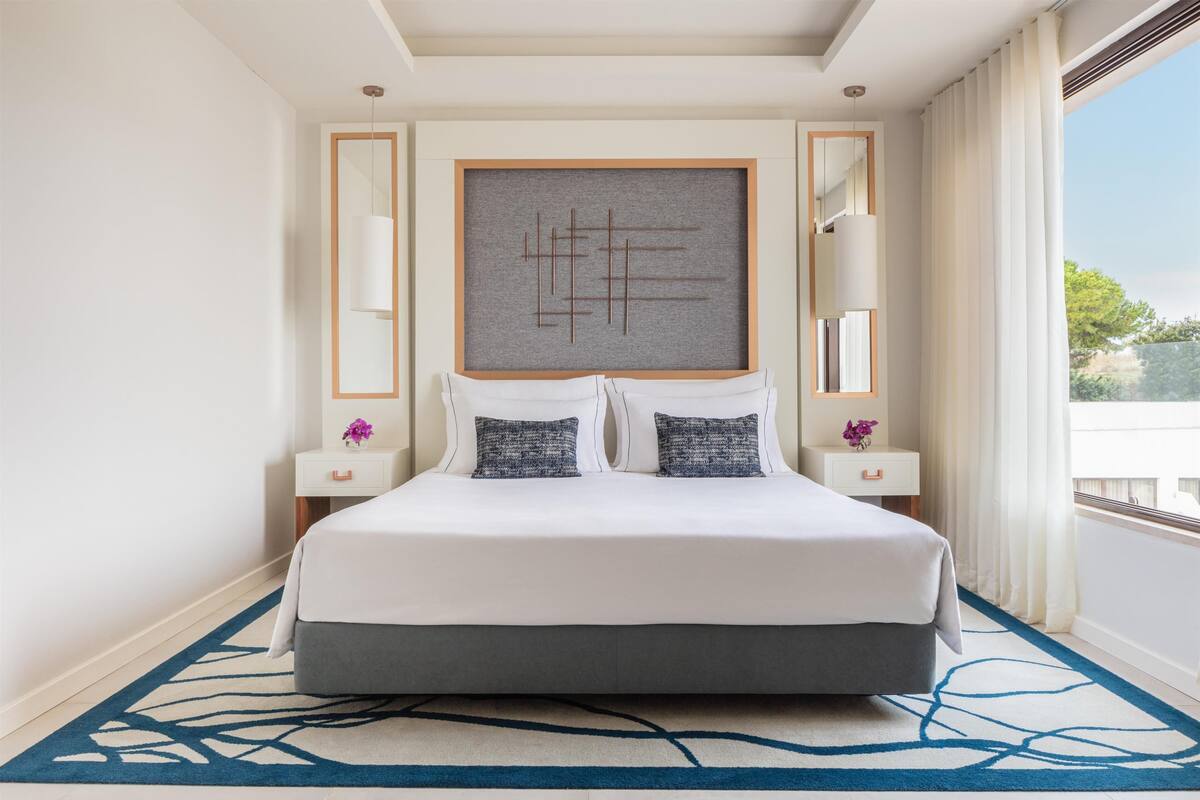 A modern hotel room with a large bed, a decorative wall piece, and a view of the outdoors.
