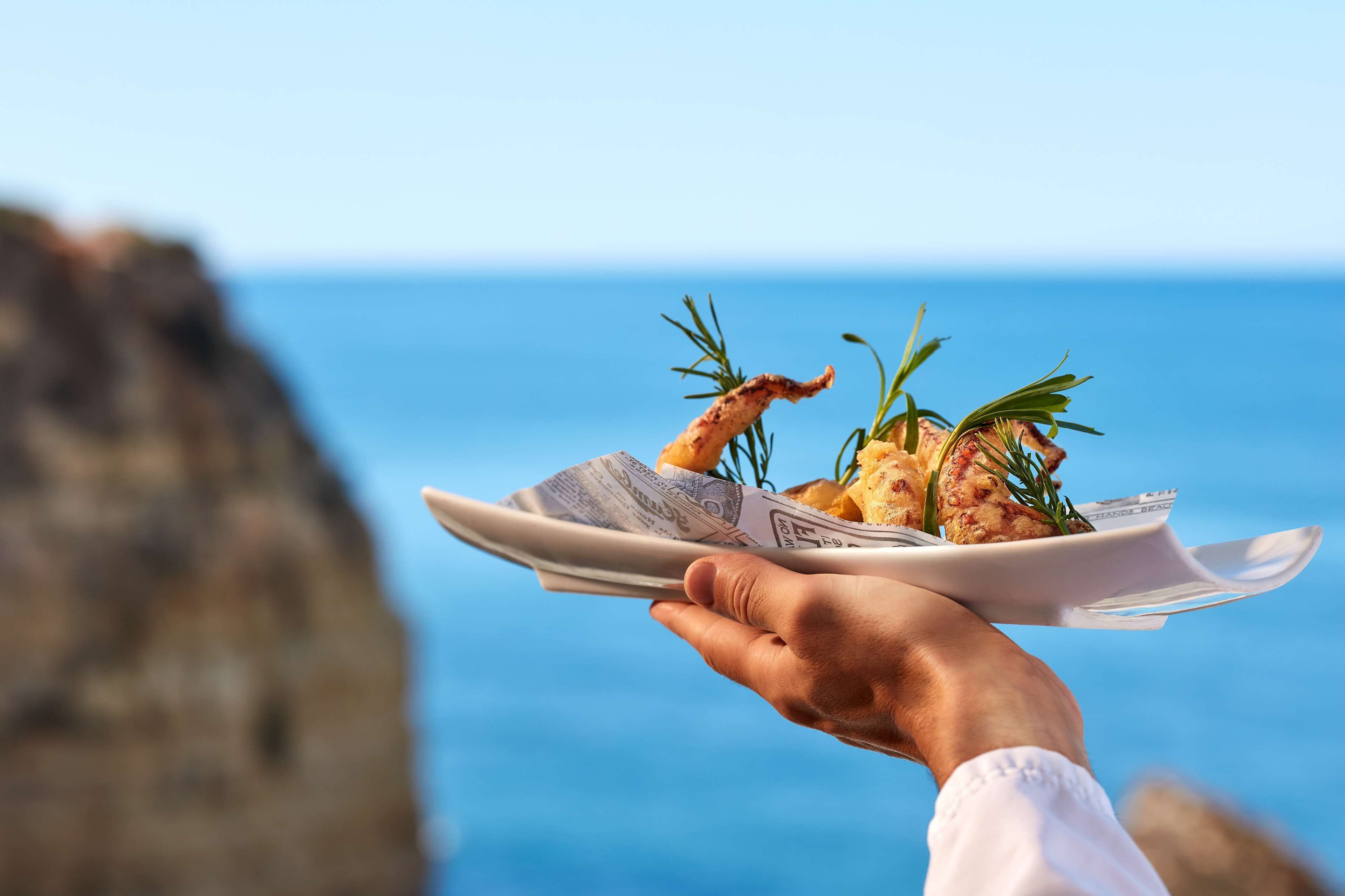 Una persona sosteniendo un plato con comida y ramitas de romero, con vista despejada al océano y acantilados rocosos al fondo.