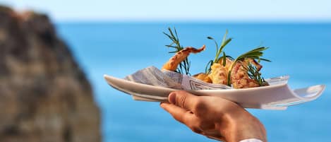 Una persona sosteniendo un plato con comida y ramitas de romero, con vista despejada al océano y acantilados rocosos al fondo.