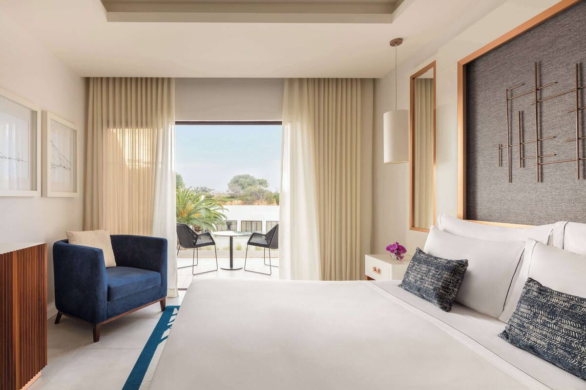 A hotel room with a large bed, a blue armchair, a small table with chairs, and a view of the outdoors through a large window.