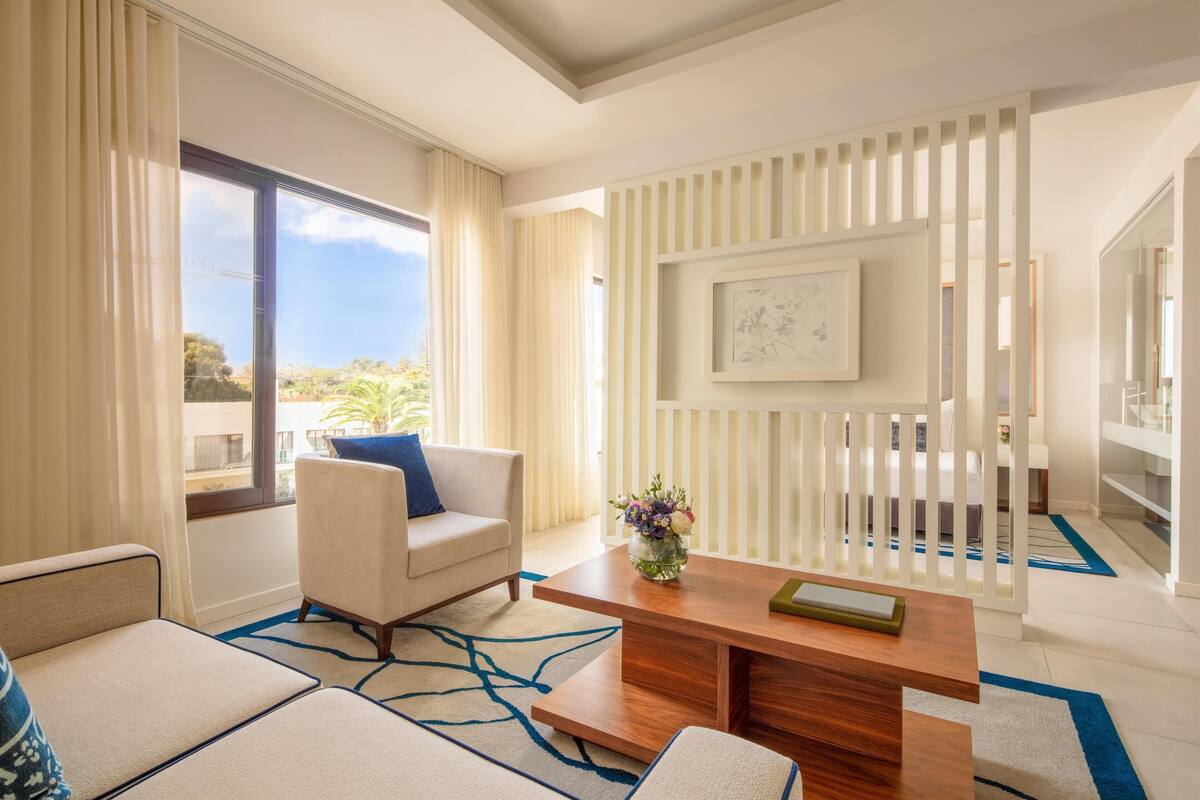 A modern hotel room with a sofa, a coffee table, and a view of the outdoors.