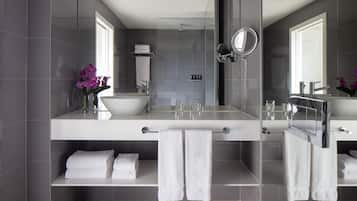 Un baño moderno con un espejo grande, dos lavamanos y una ducha. Hay toallas blancas y un florero con flores moradas en la encimera.