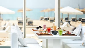 Une table à manger dressée avec différents plats et boissons, donnant sur une plage parsemée de parasols et de chaises longues.
