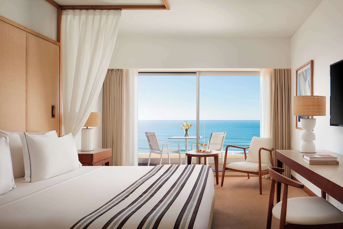 Een hotelkamer met een groot bed, een bureau met een stoel, een tafeltje met een vaas, en uitzicht op de oceaan.