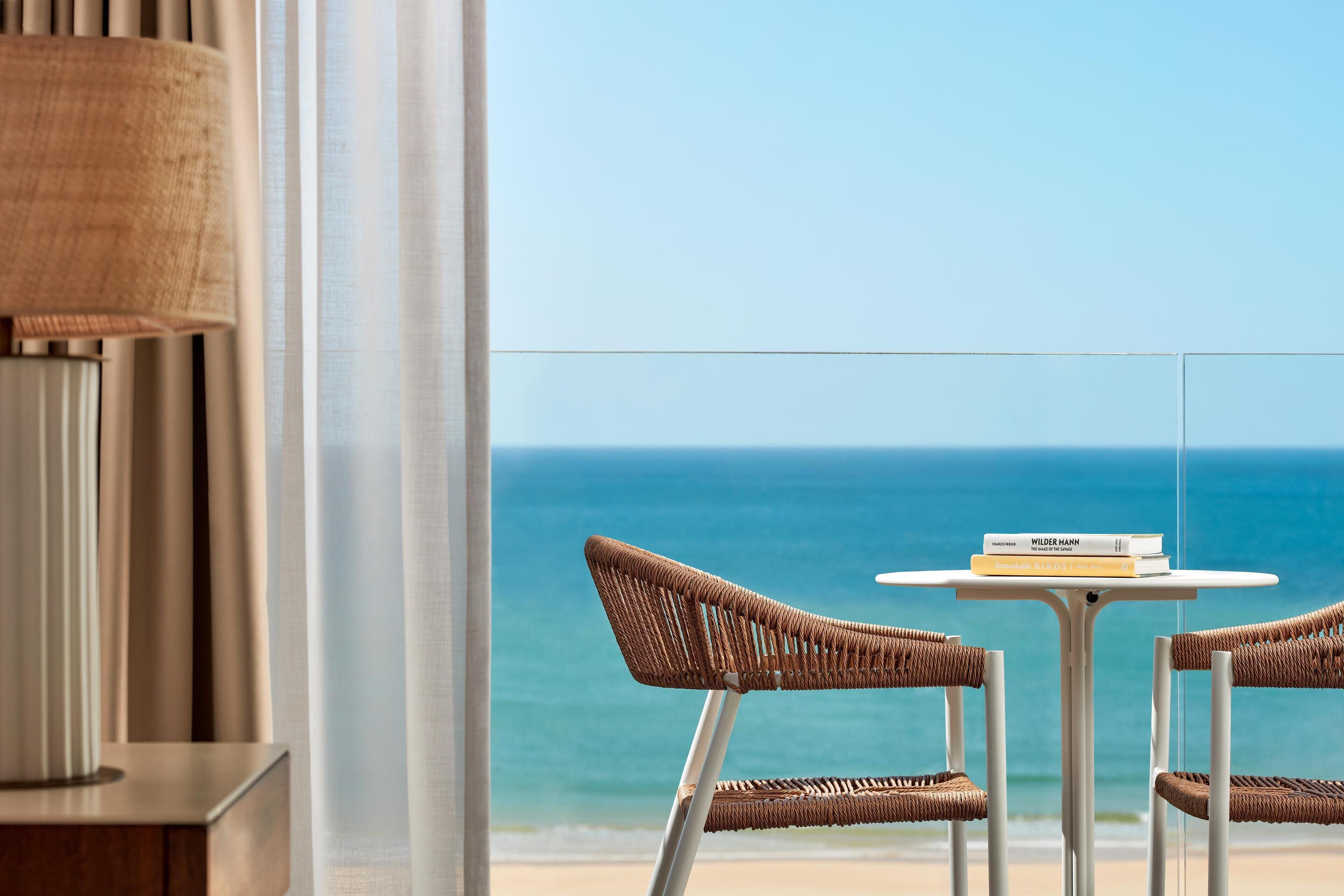 A balcony with wicker chairs and a small table, overlooking a beach and the ocean.
