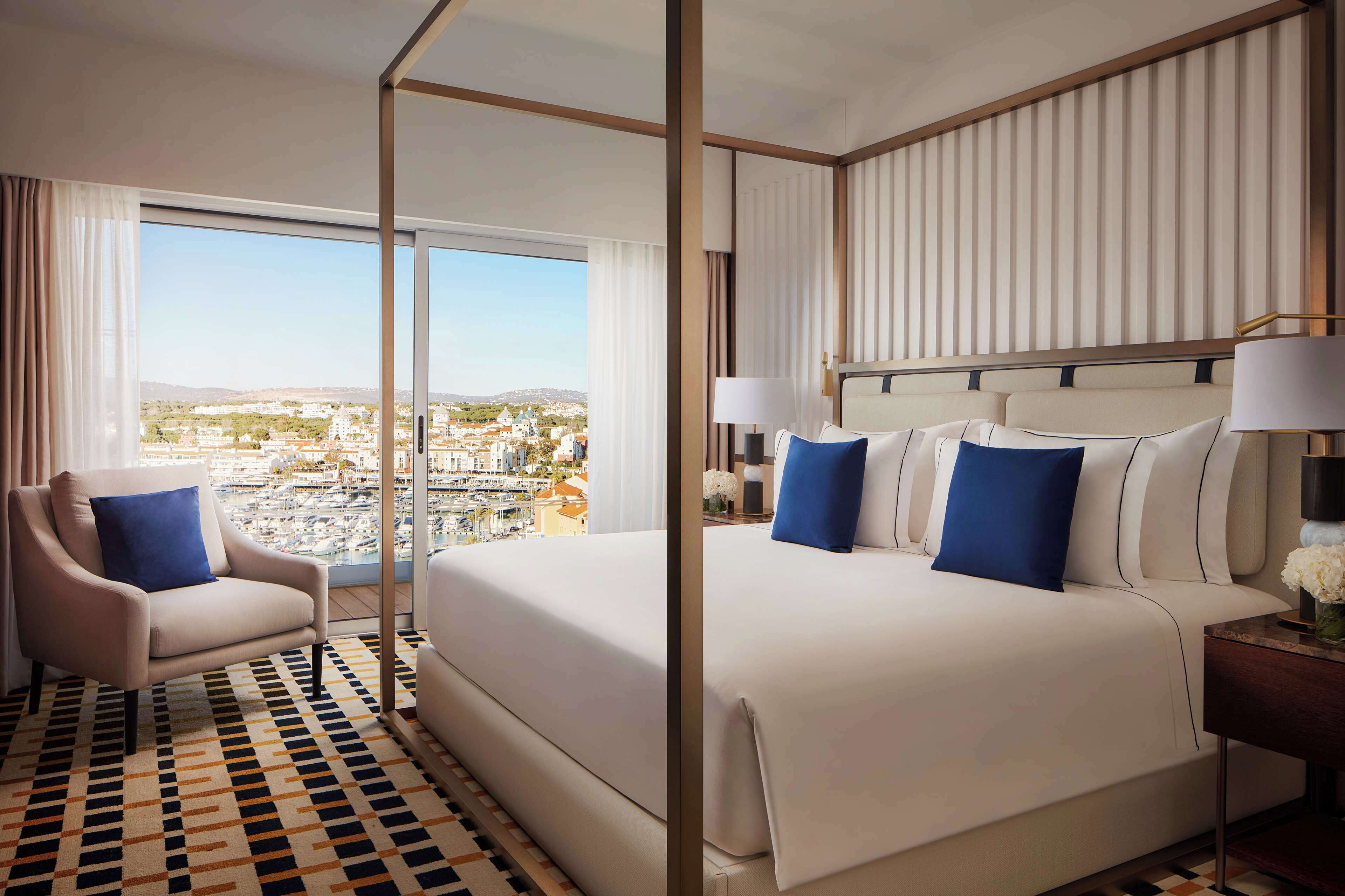 Ein Hotelzimmer mit einem großen Bett, einem Sessel, einem Nachttisch und Blick auf eine Stadtlandschaft durch große Fenster.