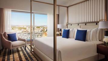 A hotel room with a large bed, a chair, a nightstand, and a view of a cityscape through large windows.