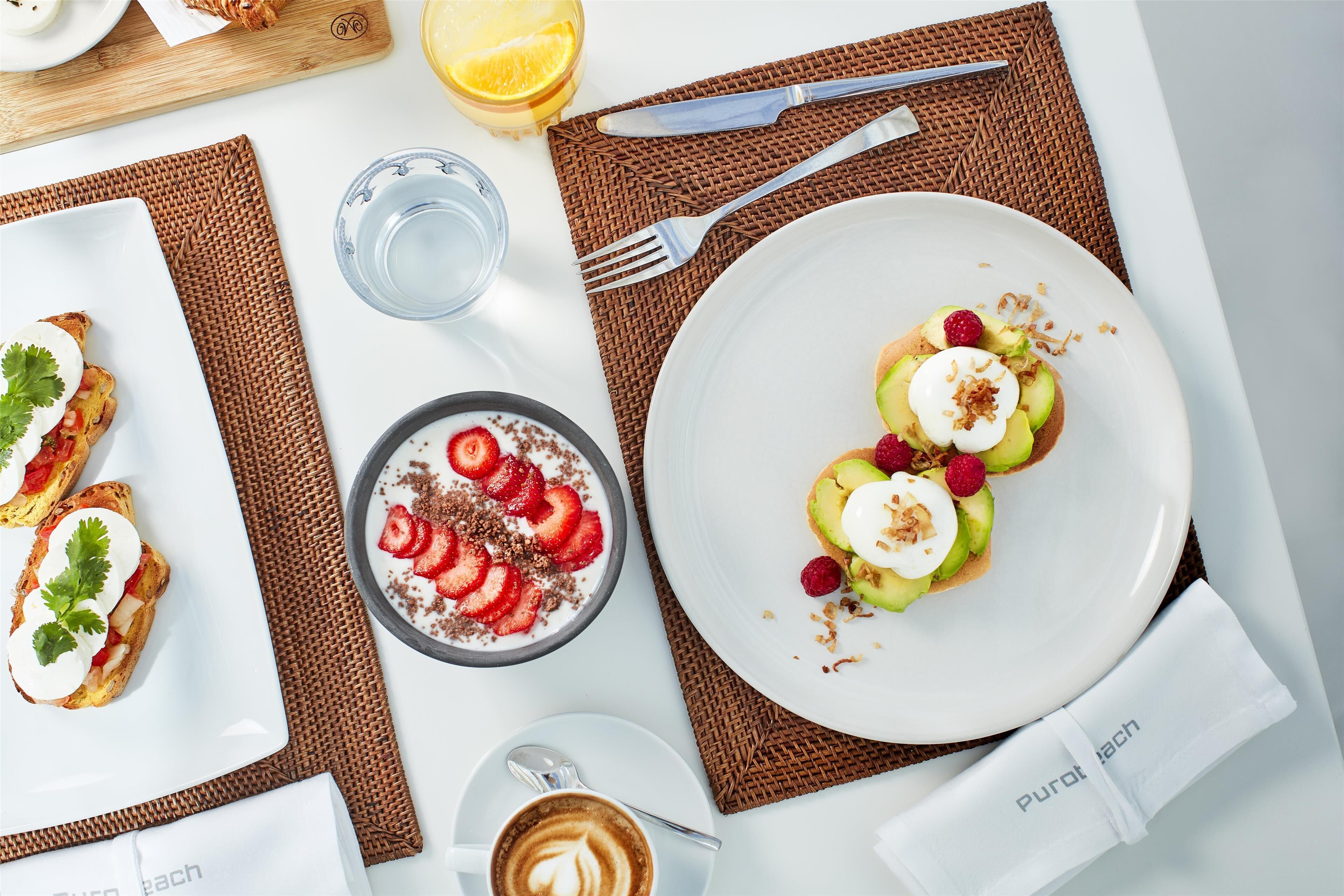 Un desayuno con tostadas, yogur, fruta y café sobre una mesa blanca.