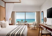 A hotel room with a large bed, a desk, a chair, a TV, and a view of the ocean.
