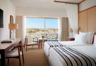A hotel room with a large bed, a desk, a chair, a small table with a vase, and a view of a cityscape through large windows.