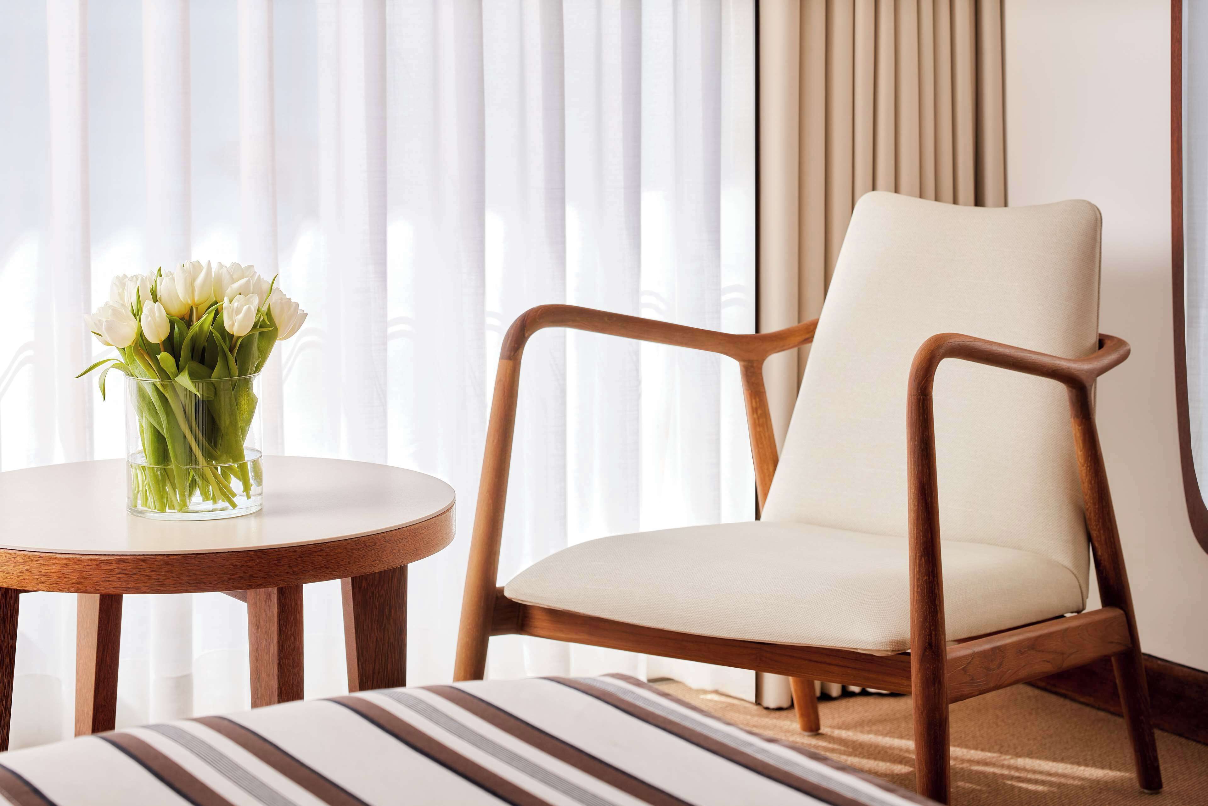 Een moderne kamer met een houten stoel en een ronde tafel, een vaas met witte bloemen en transparante gordijnen.
