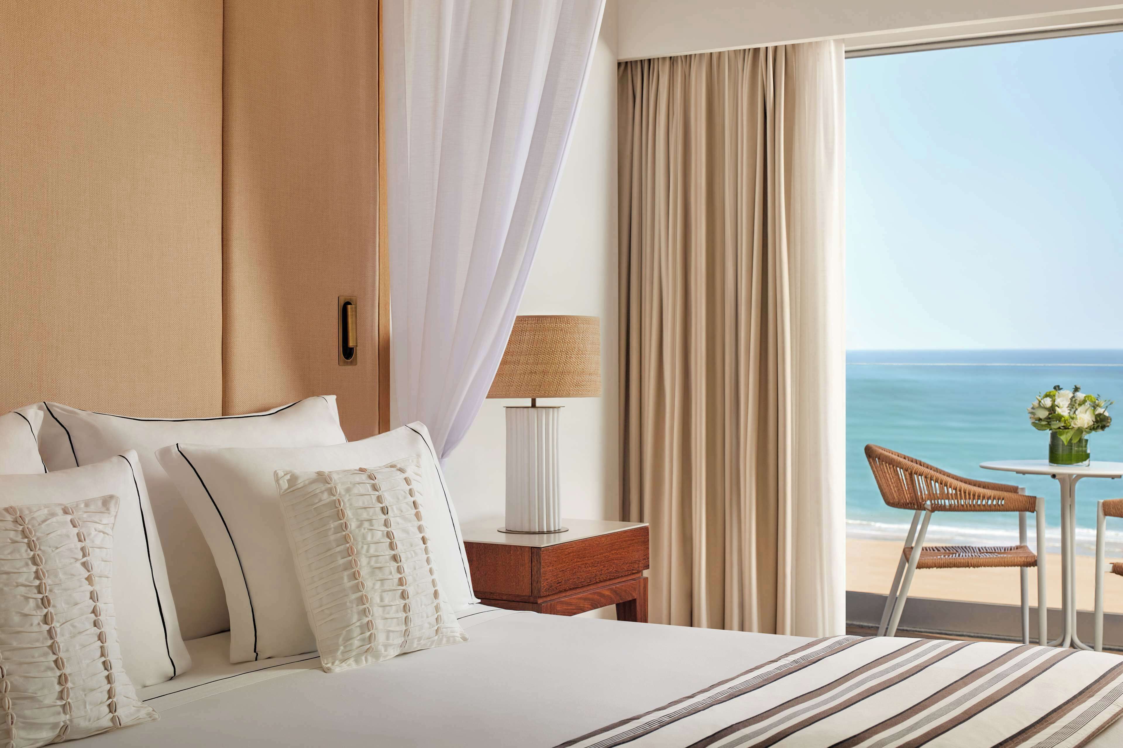 Una camera d'hotel con un letto grande, un comodino, una lampada, vista sull'oceano e un piccolo tavolo con una sedia.